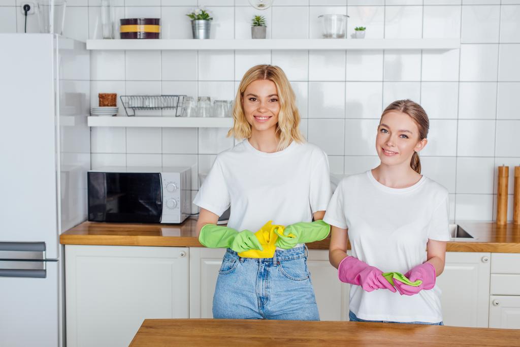 fröhliche Schwestern in Gummihandschuhen, die in der Küche in die Kamera schauen - Foto, Bild