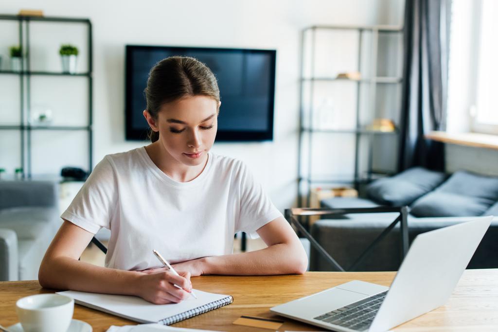 szelektív fókusz gyönyörű nő írás notebook közelében laptop és csésze kávé, online tanulmány koncepció  - Fotó, kép
