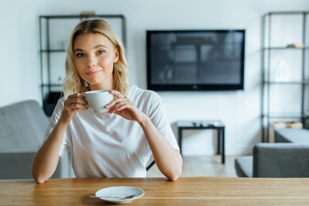 fröhliche junge Frau blickt in die Kamera und hält eine Tasse Kaffee in der Hand - Foto, Bild