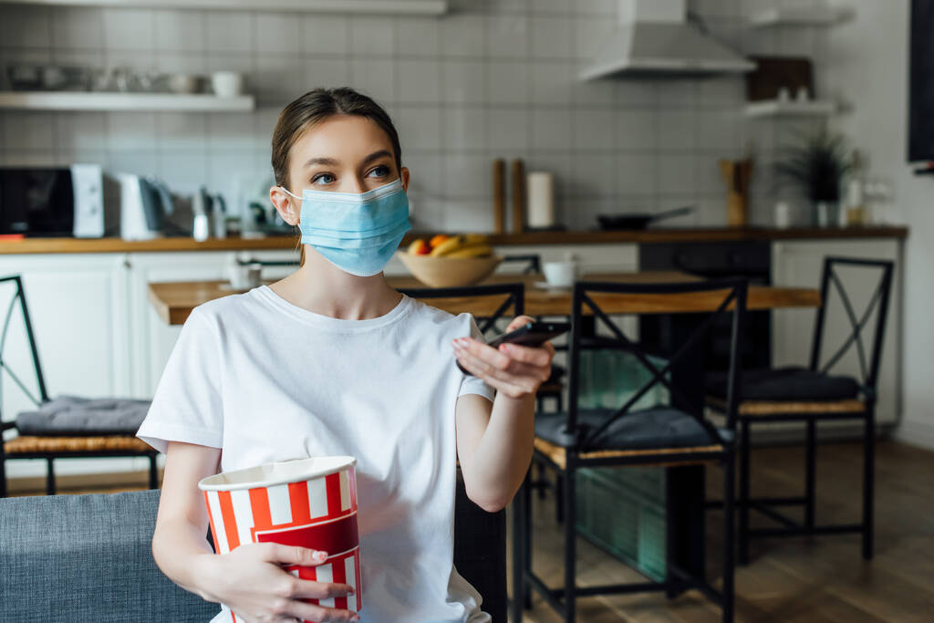 Dívka v lékařské masce drží popcorn při sledování filmu na gauči  - Fotografie, Obrázek