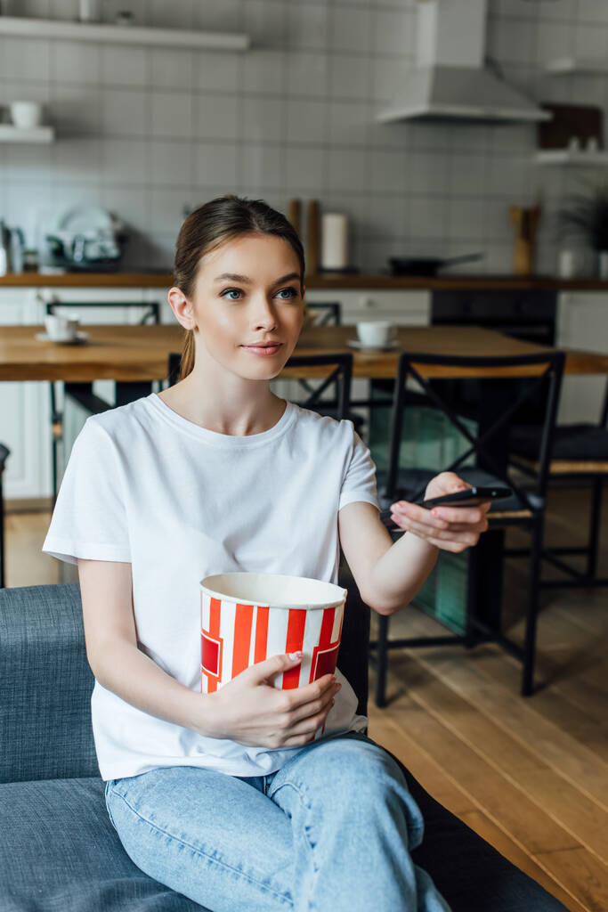 Attraente ragazza che tiene secchio di popcorn e telecomando a casa
  - Foto, immagini