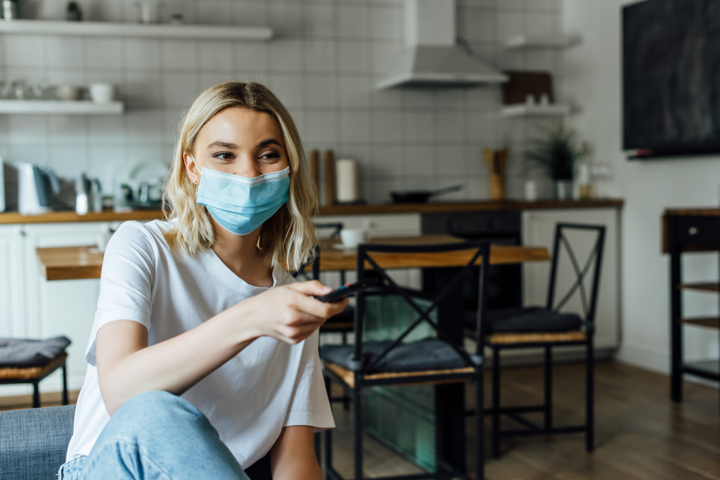 Femme blonde en masque médical tenant télécommande à la maison
  - Photo, image