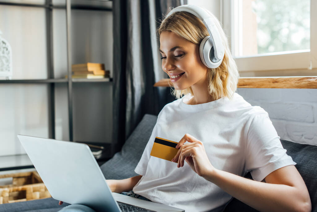 Gyönyörű mosolygós nő fülhallgató kezében hitelkártya és laptop a kanapén  - Fotó, kép