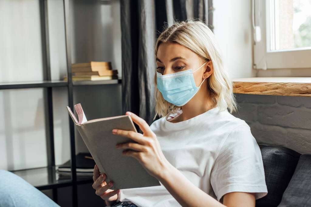 Blond vrouw in medisch masker lezen boek thuis  - Foto, afbeelding