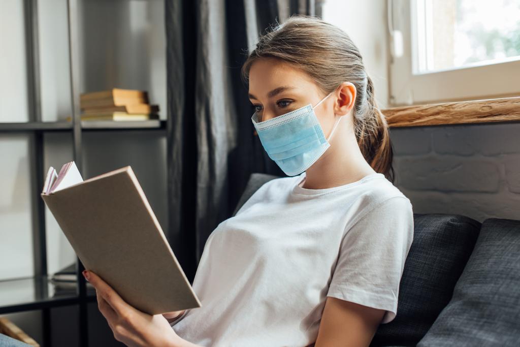 Νεαρή γυναίκα σε ιατρική μάσκα βιβλίο ανάγνωσης στον καναπέ  - Φωτογραφία, εικόνα
