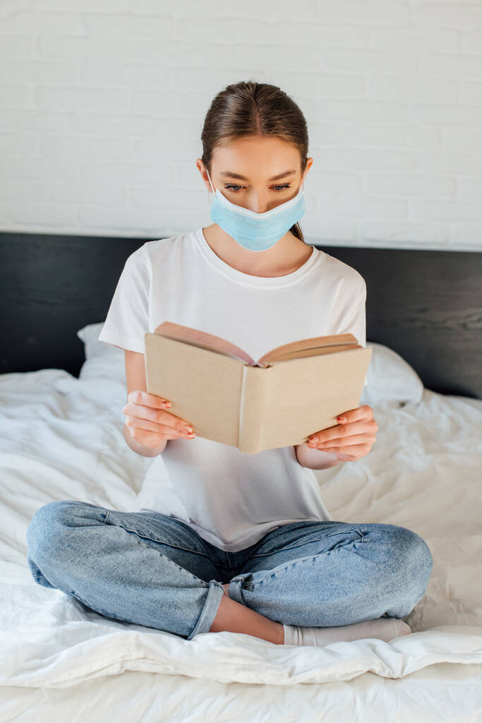Mladá žena v lékařské masce čtení knihy, zatímco sedí se zkříženýma nohama na posteli  - Fotografie, Obrázek