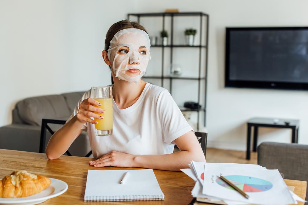 Focus selettivo della donna in maschera che tiene il bicchiere di succo d'arancia vicino a carte con grafici sul tavolo
  - Foto, immagini