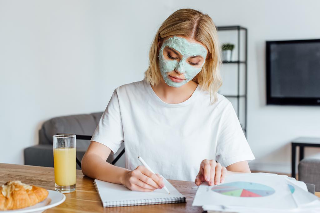 Valikoiva painopiste blondi nainen kasvot naamio työskentelee asiakirjoja ja muistikirja lähellä croissant ja appelsiinimehu pöydällä
  - Valokuva, kuva