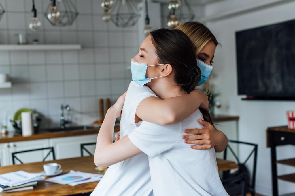 Widok z boku sióstr w maskach medycznych przytulających się w domu  - Zdjęcie, obraz
