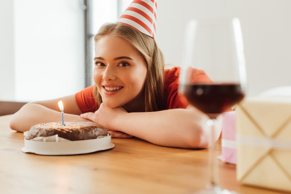 selective focus of happy girl in party cap looking at camera near birthday cake, presents and wine glass on table - Photo, Image
