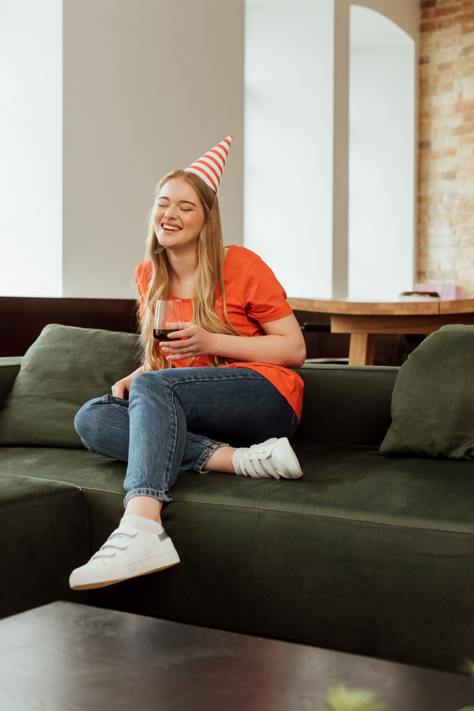 вибірковий фокус щасливої жінки в партійній кепці, сидячи на дивані і тримаючи келих червоного вина
  - Фото, зображення