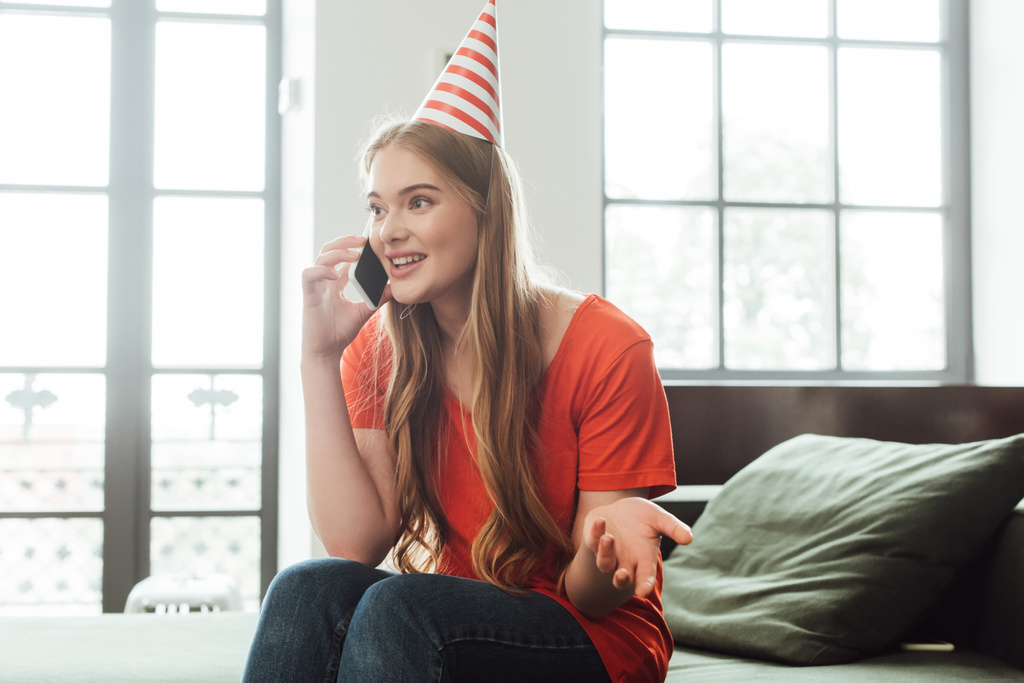 веселая девушка в кепке для вечеринок жестикулирует и разговаривает по смартфону дома
  - Фото, изображение