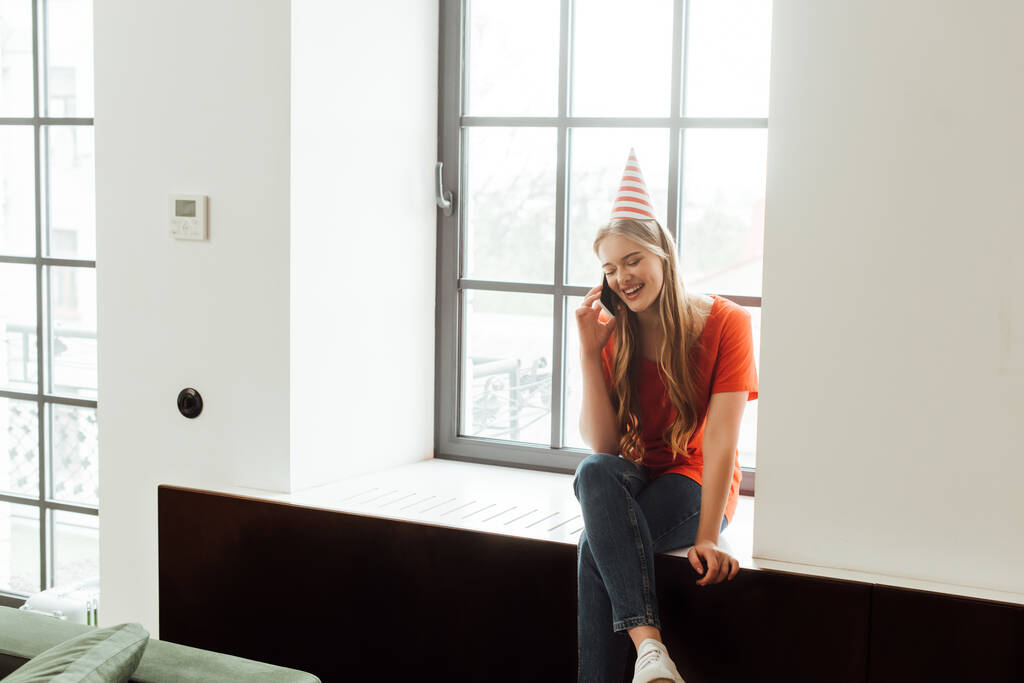 улыбающаяся девушка в кепке для вечеринок сидит на подоконнике и разговаривает по смартфону
 - Фото, изображение