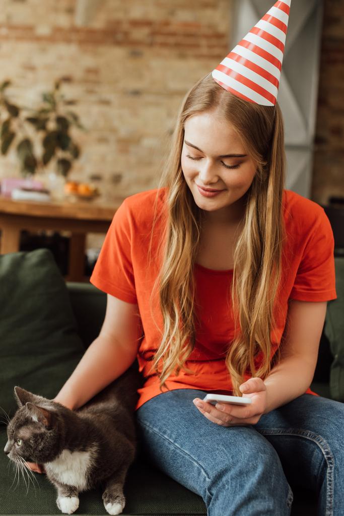 Parti şapkalı neşeli genç kadın sevimli kediye dokunuyor ve akıllı telefon tutuyor. - Fotoğraf, Görsel