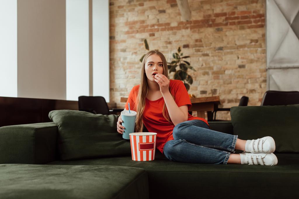 aantrekkelijk meisje eten popcorn, holding wegwerpbeker en het kijken naar film in de woonkamer  - Foto, afbeelding