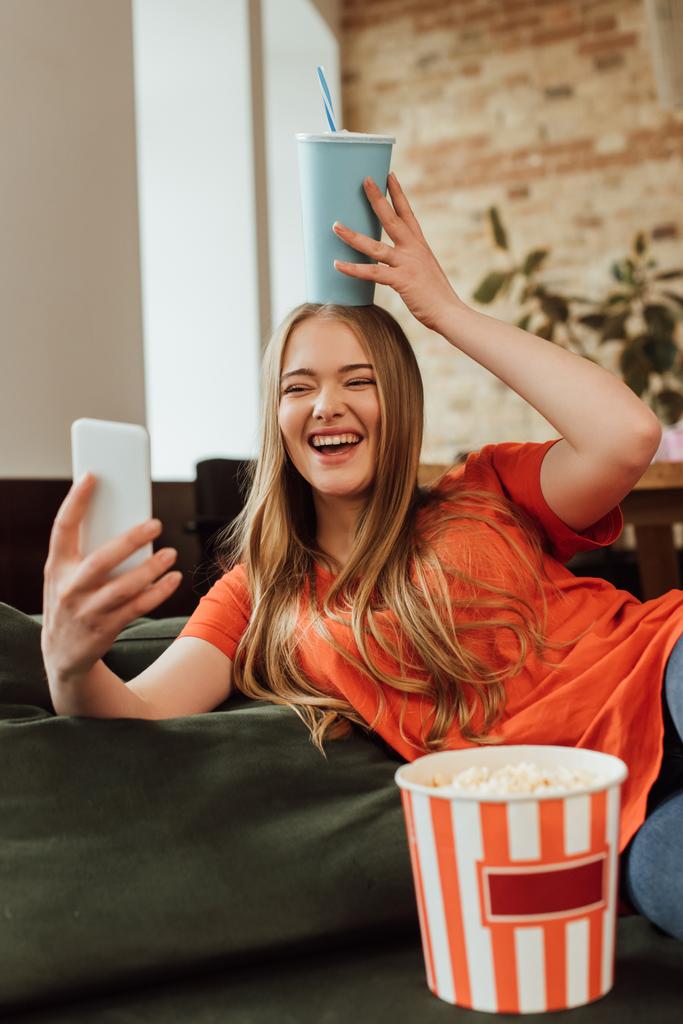 selectieve focus van vrolijk meisje houden papieren beker boven het hoofd en het nemen van selfie in de buurt van popcorn emmer  - Foto, afbeelding