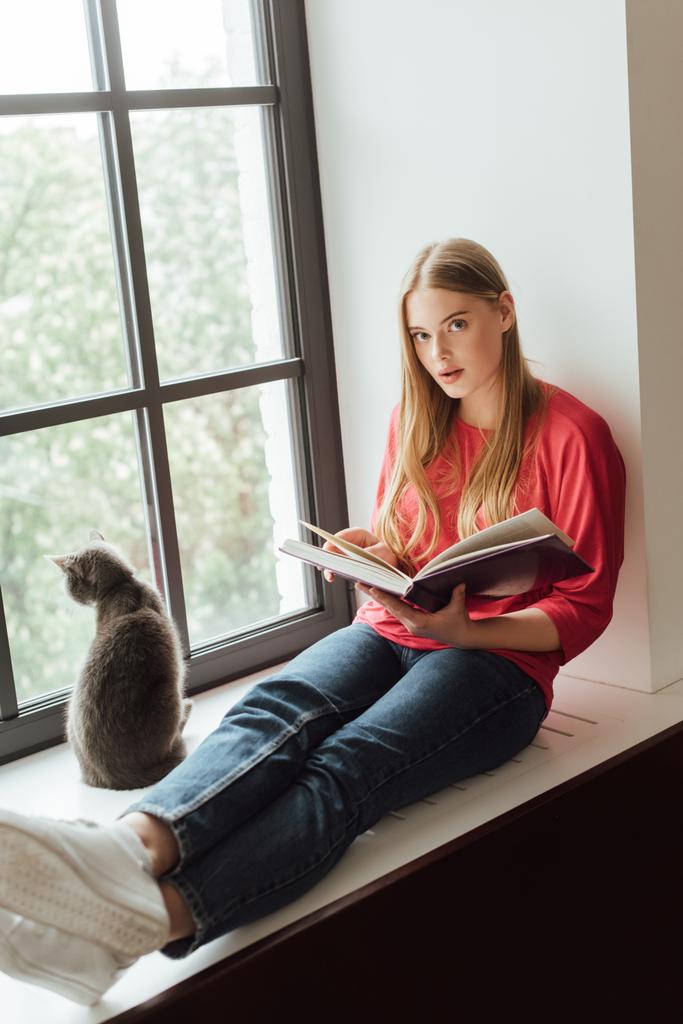 Güzel kız kitap tutuyor ve pencerenin kenarında sevimli bir kedinin yanında oturuyor.  - Fotoğraf, Görsel
