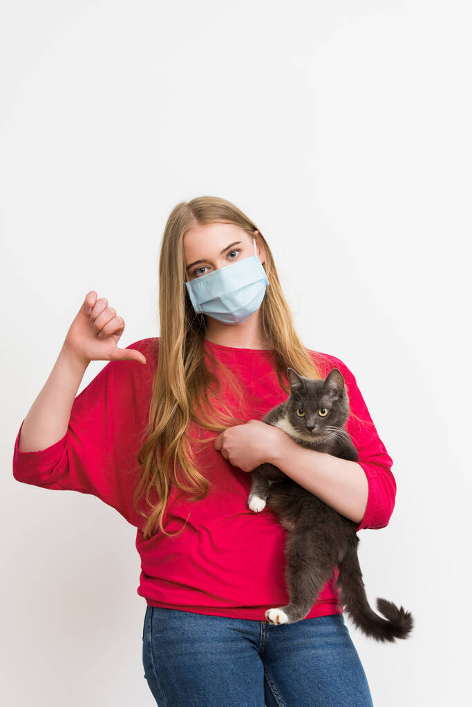 mladá žena v lékařské masce držení v náručí roztomilé kočky a ukazující palec dolů izolované na bílé  - Fotografie, Obrázek