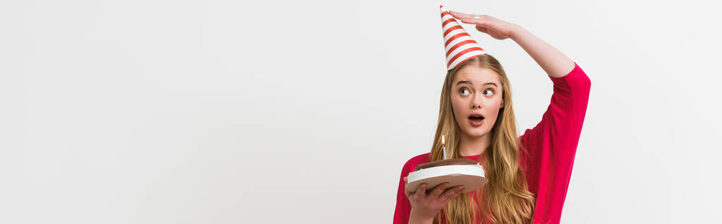 панорамна концепція здивованої дівчини торкається вечірки кепка і тримає торт на день народження ізольовано на білому
  - Фото, зображення