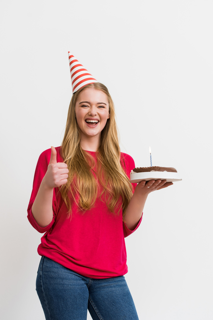 Parti şapkalı neşeli kız baş parmağını gösteriyor ve beyaz üzerinde izole edilmiş doğum günü pastası tutuyor.  - Fotoğraf, Görsel