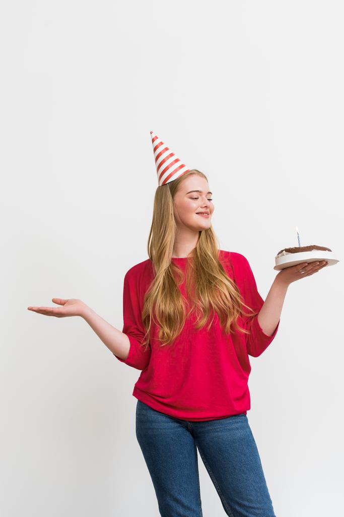 νεαρή γυναίκα με καπέλο κόμμα κρατώντας τούρτα γενεθλίων και gesturing απομονώνονται σε λευκό  - Φωτογραφία, εικόνα