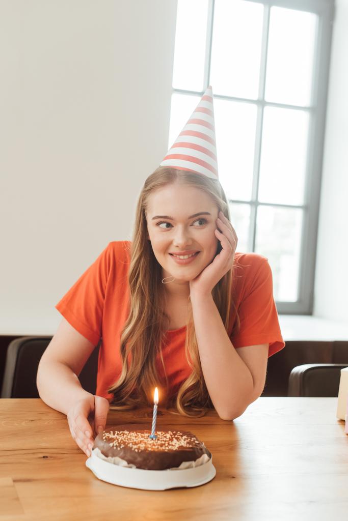Χαρούμενη γυναίκα με καπέλο κόμμα κοιτάζοντας μακριά κοντά νόστιμα τούρτα γενεθλίων  - Φωτογραφία, εικόνα