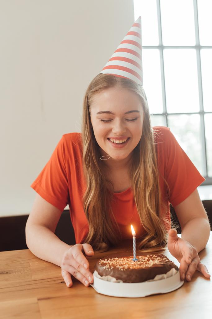 wesoła kobieta w party cap patrząc na pyszne tort urodzinowy  - Zdjęcie, obraz