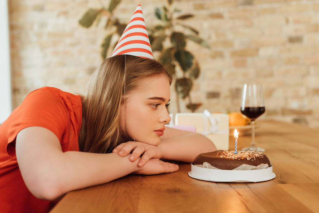 schönes Mädchen in Partymütze schaut auf leckeren Geburtstagskuchen neben Weinglas und Geschenke auf dem Tisch - Foto, Bild