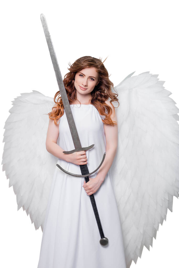 Μια νεαρή γυναίκα, ένας άγγελος με φτερά σε ένα λευκό φόρεμα με ιπτάμενα μαλλιά ποζάρουν με ένα σπαθί στα χέρια του απομονώνονται σε λευκό φόντο. - Φωτογραφία, εικόνα