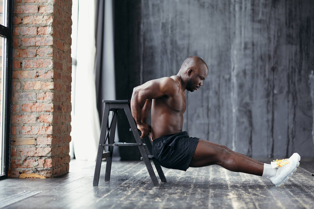 Profile view of a sporty African American with naked torso and sneakers leans on chair and shakes his arm muscles - Photo, Image
