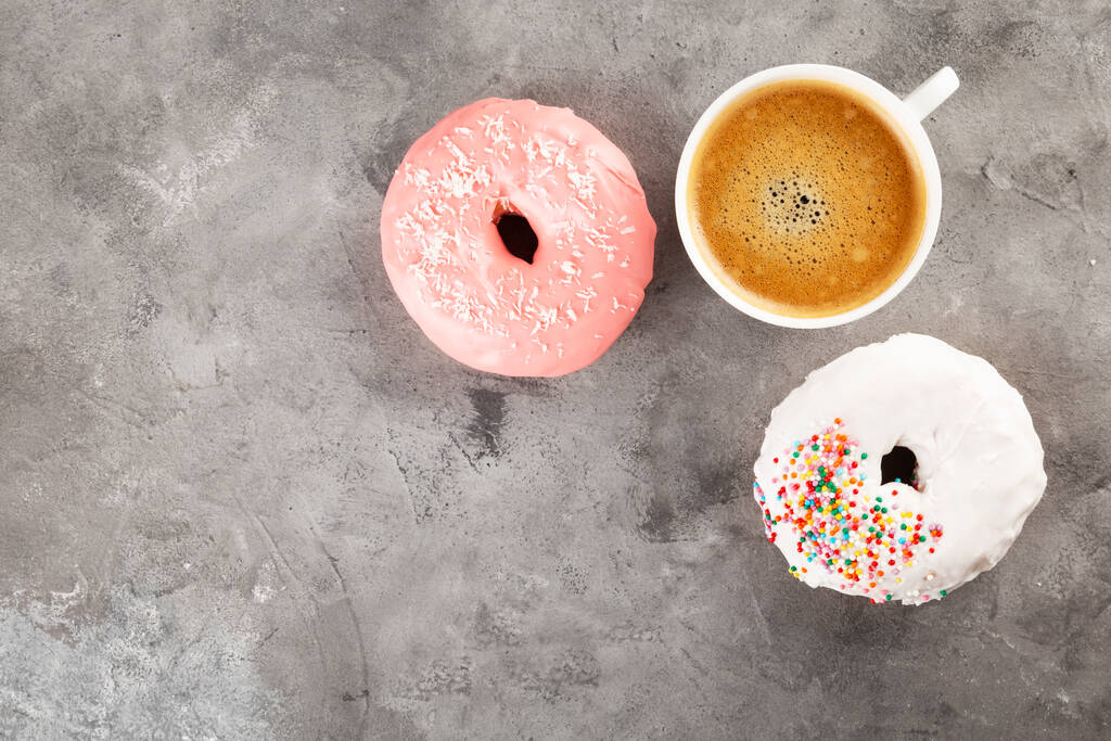 白とピンクのアイシングとグレーの背景にトッピングされた多色の砂糖菓子でコーヒーとドーナツの白いカップ。最上階だ。スペースのコピー - 写真・画像