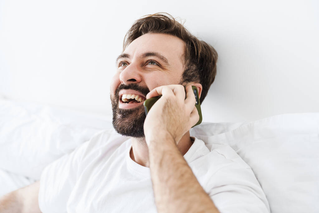 Imagem do jovem homem caucasiano não barbeado sorrindo e falando no celular enquanto estava deitado na cama em casa
 - Foto, Imagem