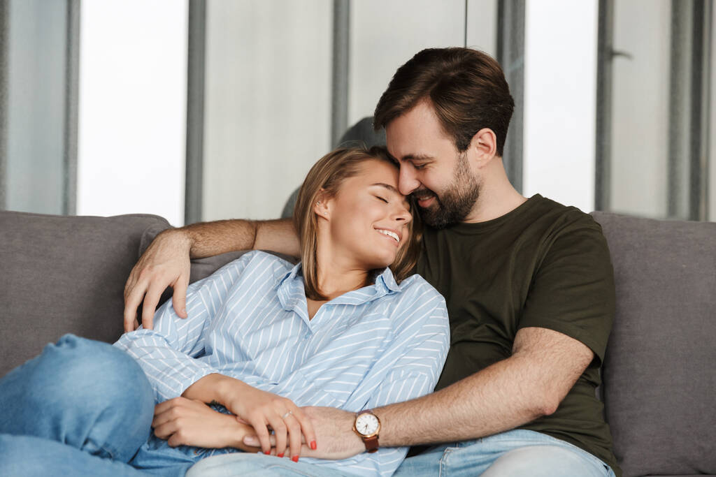 Фото веселой молодой пары, улыбающейся и обнимающейся, сидя дома на диване
 - Фото, изображение