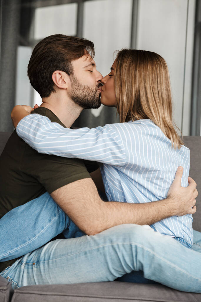 Foto de casal romântico feliz caucasiano beijando e abraçando enquanto sentado no sofá em casa
 - Foto, Imagem