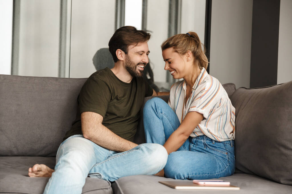 Foto van vrolijke blanke echtpaar lachen en praten terwijl zitten op de bank thuis - Foto, afbeelding