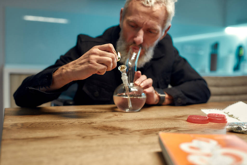 Seeking relaxation. Senior man lighting cannabis in the bowl of glass water pipe or bong in the kitchen. Marijuana tools on the table. Legalization and dependence on light drugs concept - Photo, Image