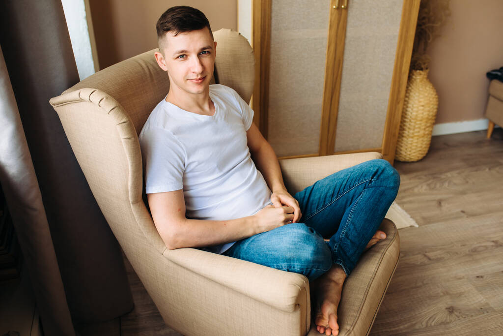 Egy fiatal fickó ül egy bézs fotelben mezítláb és a kamerába néz. Alkalmi stílus - Fotó, kép