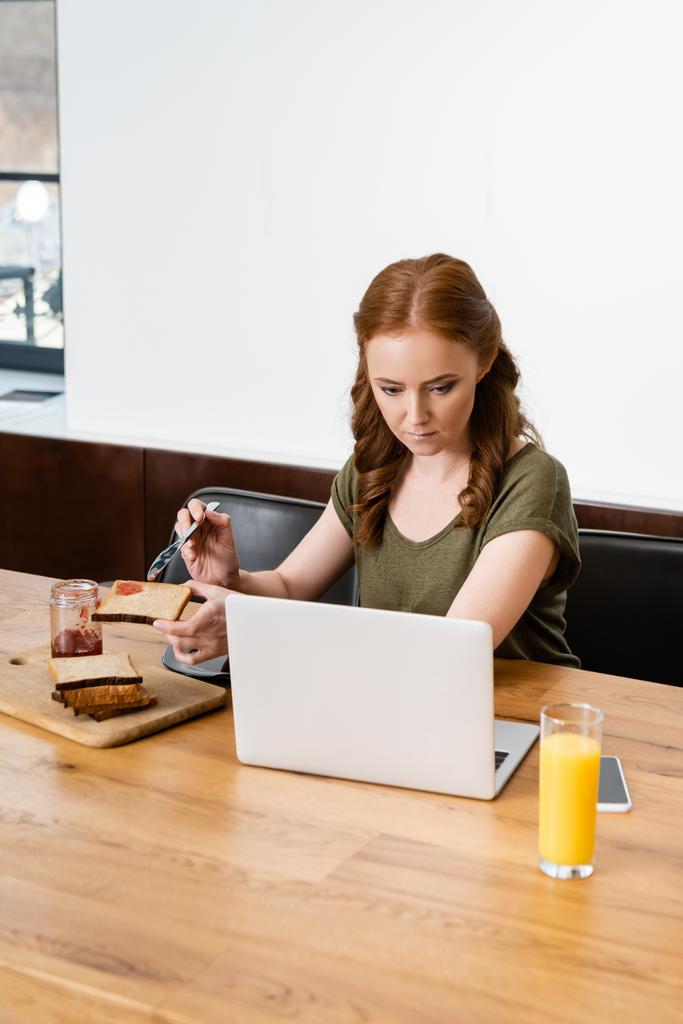 A laptopot nézegető nő szelektív fókusza, miközben a pirítóst lekvárral tartja az asztalnál  - Fotó, kép