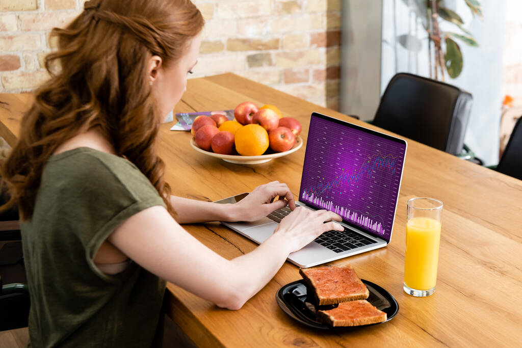 Vista lateral da mulher usando laptop com gráficos perto de torradas e suco de laranja na mesa
  - Foto, Imagem