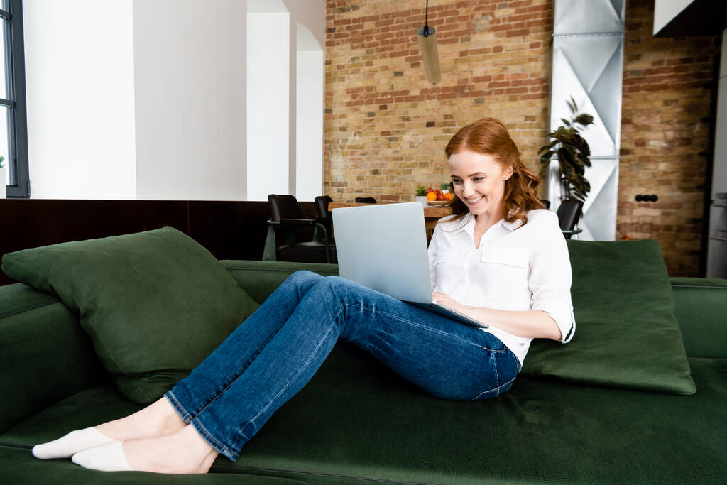Красивий телефонний працівник посміхається, використовуючи ноутбук на дивані вдома
  - Фото, зображення