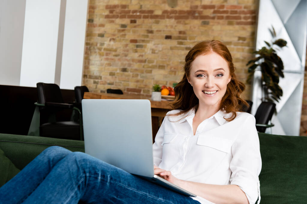 Selectieve focus van mooie vrouw glimlachend op de camera tijdens het gebruik van laptop op de bank  - Foto, afbeelding
