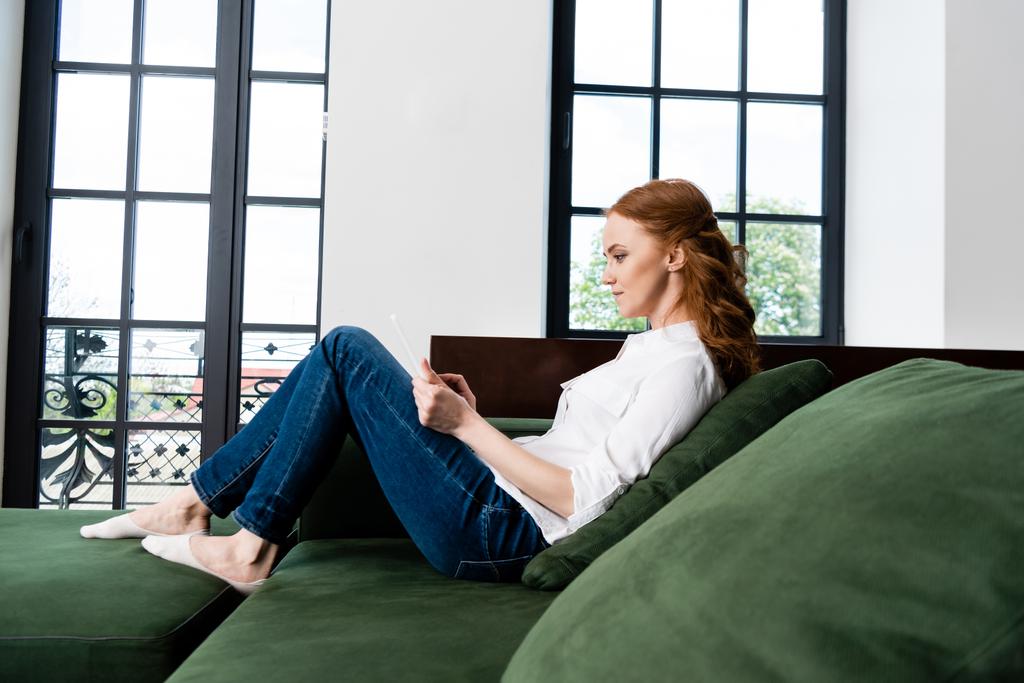 Seitenansicht der schönen rothaarigen Frau mit digitalem Tablet auf dem Sofa  - Foto, Bild
