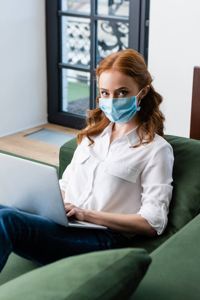 Foco seletivo da mulher de cabelos vermelhos em máscara médica olhando para a câmera ao usar laptop no sofá
  - Foto, Imagem