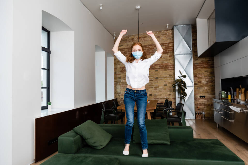 Woman in medical mask showing yeah gesture while jumping on couch  - Photo, Image