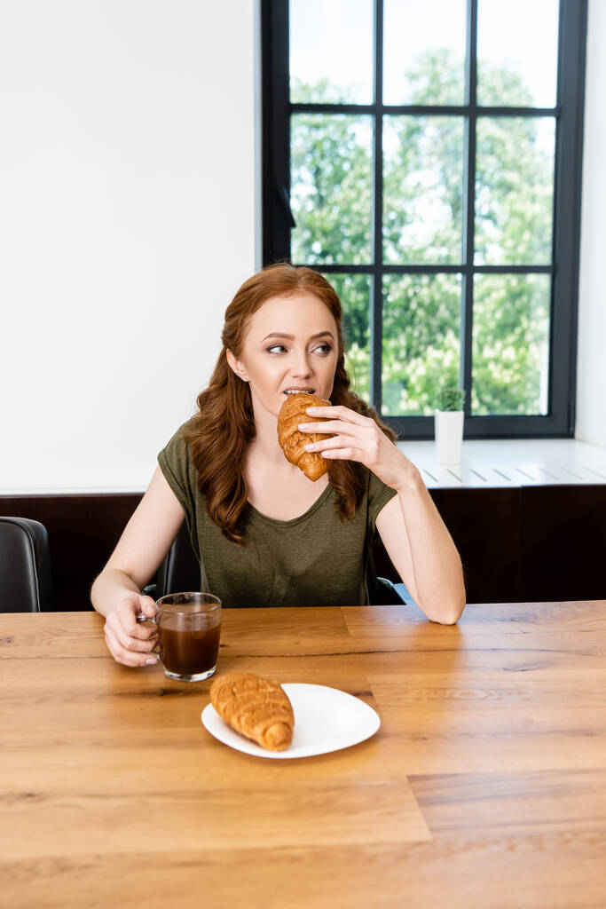 Επιλεκτική εστίαση της γυναίκας τρώει κρουασάν και κρατώντας φλιτζάνι καφέ στο τραπέζι  - Φωτογραφία, εικόνα