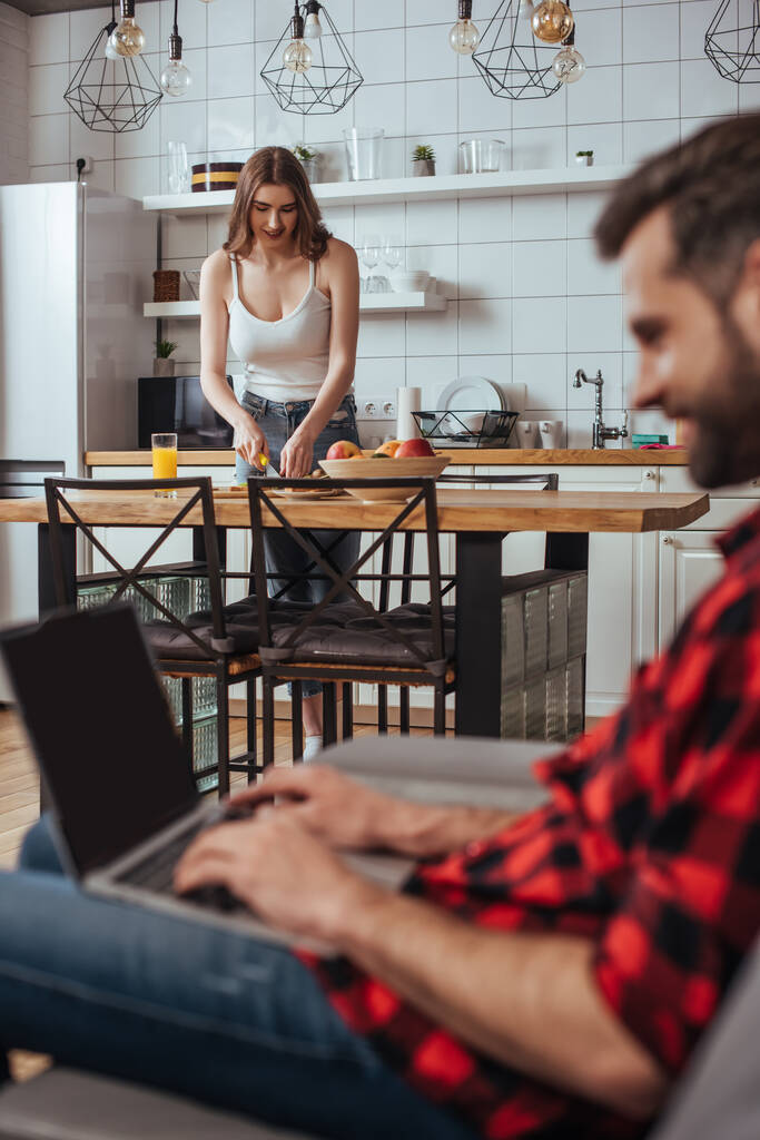 selective focus of freelancer working on laptop with blank screen in kitchen near attractive girlfriend on background - Photo, Image