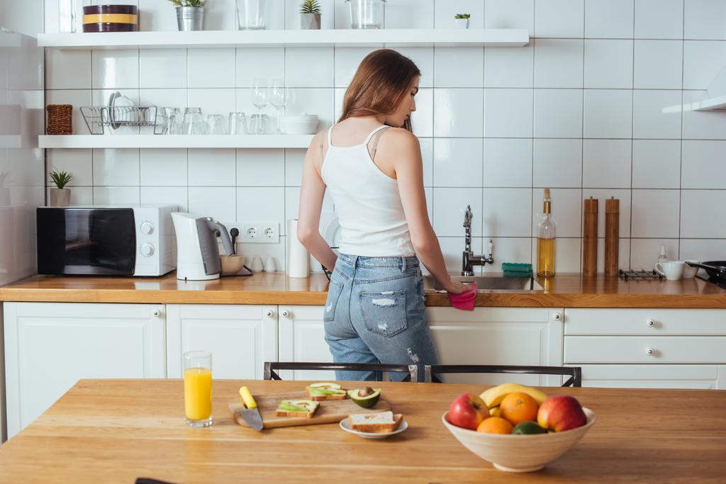 Вид сзади молодой женщины, готовящей завтрак на современной кухне рядом со столом со свежими фруктами
 - Фото, изображение