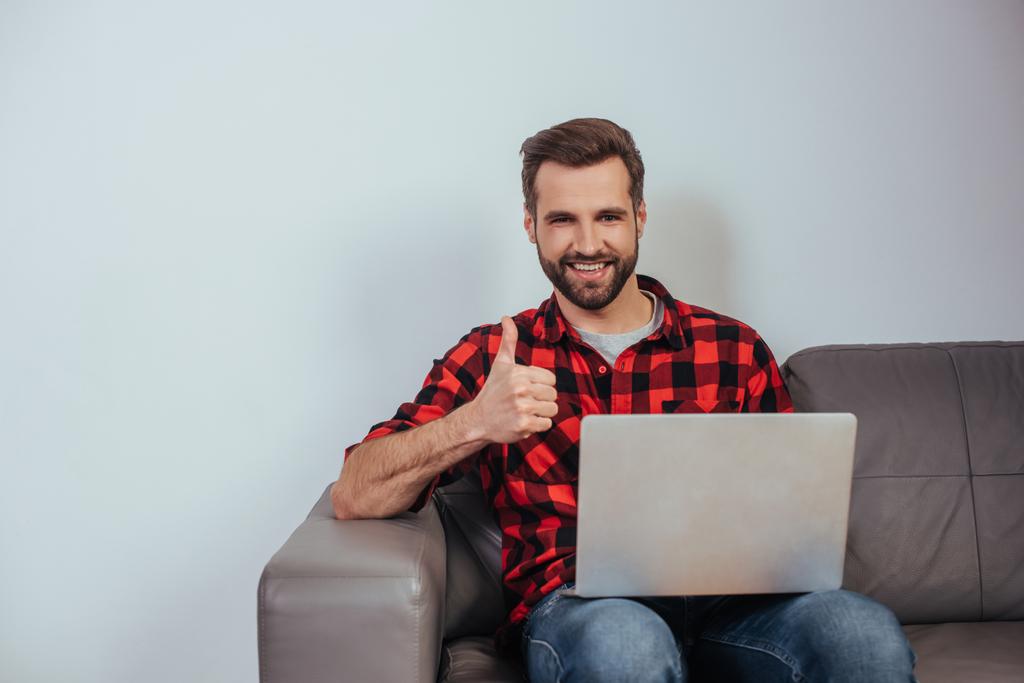 happy freelancer smiling at camera and showing thumb up while sitting on sofa with laptop - Photo, Image