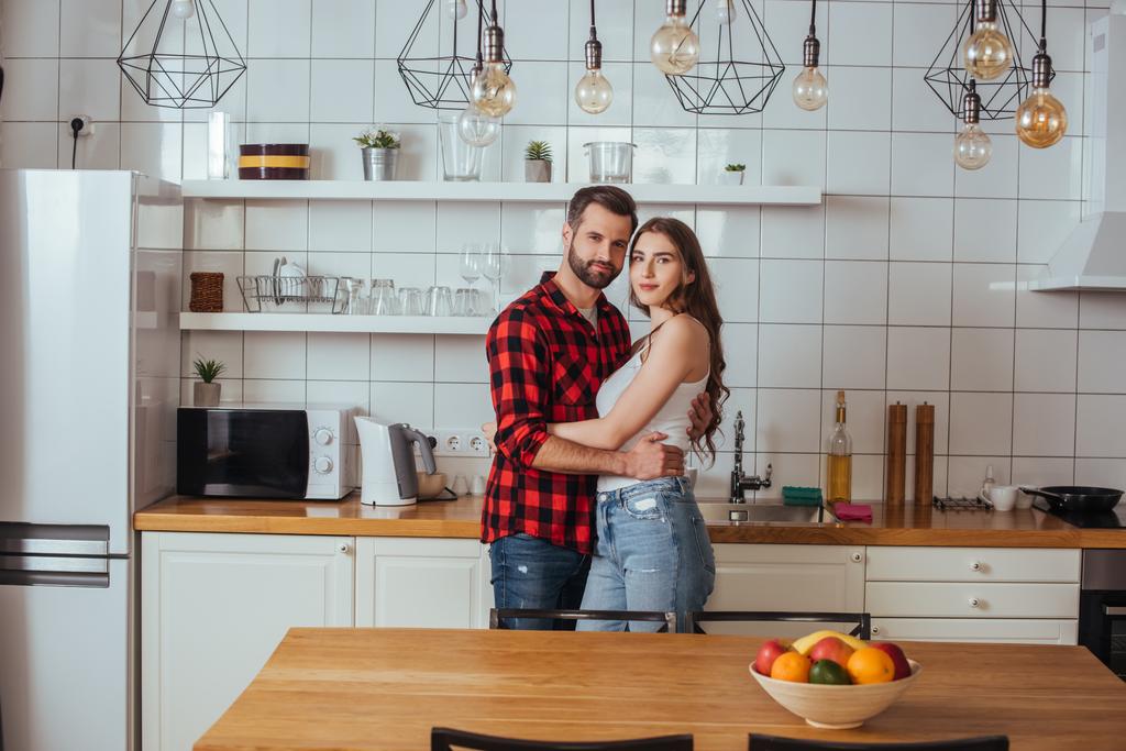 heureux jeune couple embrasser et regarder la caméra tout en se tenant dans la cuisine moderne - Photo, image