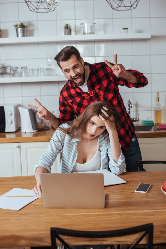 vidám férfi bolondozik körül kimerült barátnő ül mellett laptop és notebook a konyhában - Fotó, kép